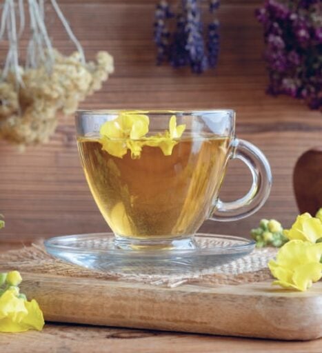 Tisane aux fleurs dans une tasse en verre, idéale pour la relaxation, avec des fleurs jaunes flottant à la surface, sur une planche en bois, décorée de fleurs séchées violettes et jaunes, ambiance apa.