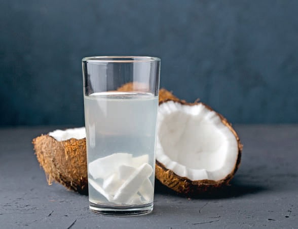 Verre d'eau avec morceaux de noix de coco et coquille de noix de coco fraîche en arrière-plan, sur fond gris foncé.