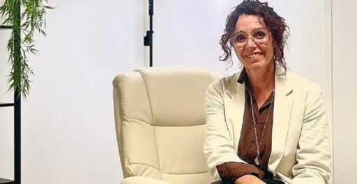 Femme professionnelle assise dans un bureau moderne, souriante, portant une veste beige, avec un fauteuil ergonomique, symbolisant le bien-être et la santé.