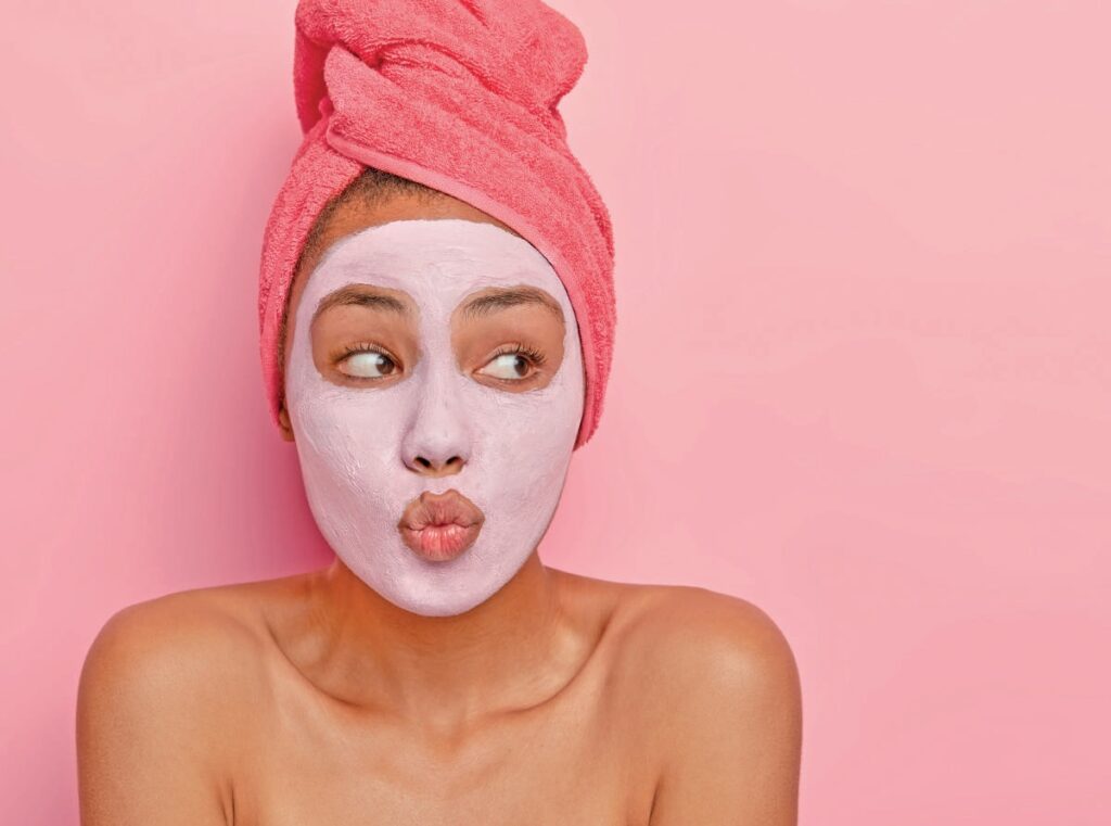 Jeune femme avec masque facial blanc, serviette rose sur la tête, exprimant une surprise ou une curiosité, contre un fond rose pastel, illustrant les soins de beauté et de bien-être.