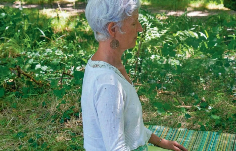 Femme âgée méditant en position de yoga dans un environnement naturel verdoyant, symbolisant la détente et le bien-être.