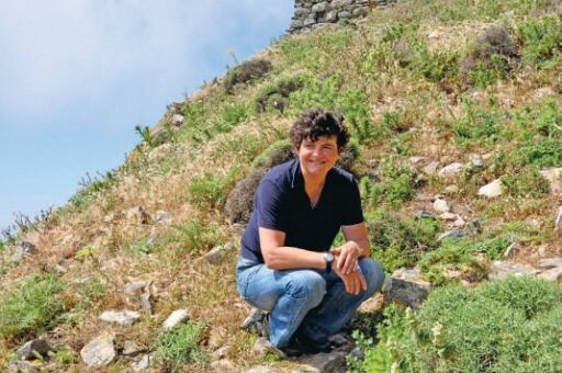 Femme souriante en pleine nature, assise sur un terrain en pente avec des plantes et des rochers, illustrant un moment de détente et de réflexion dans un environnement naturel.