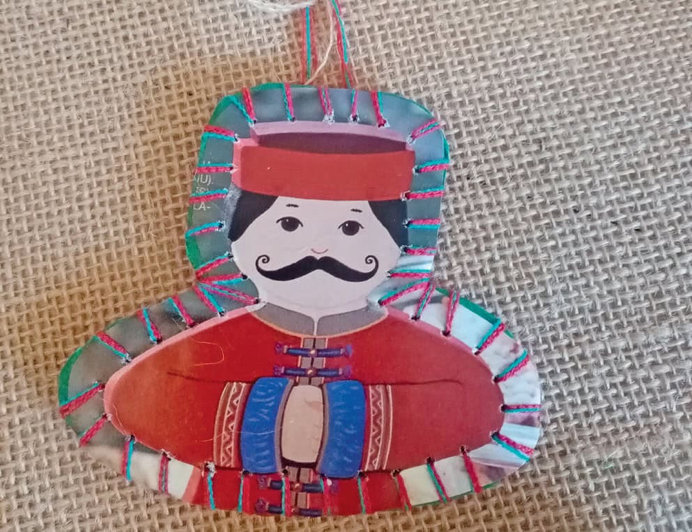 1. Marionnette en papier colorée représentant un cavalier avec chapeau et moustache, décorée de fils multicolores, posée sur une toile de jute.