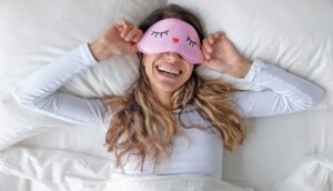Femme souriante portant un masque de sommeil rose avec des cils et un cœur rouge, allongée dans un lit blanc, évoquant relaxation et bien-être.