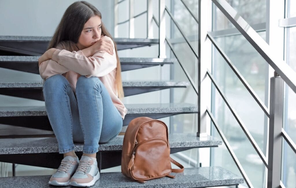 Jeune femme assise sur un escalier d'aéroport, l'air préoccupé, avec un sac à dos en cuir à côté d'elle, illustrant l'anxiété ou le stress liés aux voyages.